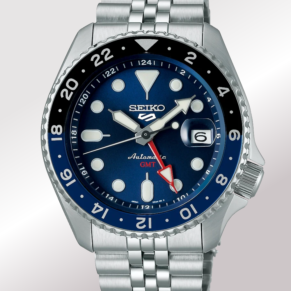 A Seiko 5 sports Automatic GMT blueberry watch with a silver stainless steel case and bracelet, featuring a deep blue dial, a black rotating bezel, and white, red, and blue accents.